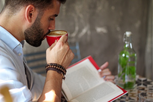 A bearded man sips coffee while reading a self-improvement book, which is one example of a professional development goal.