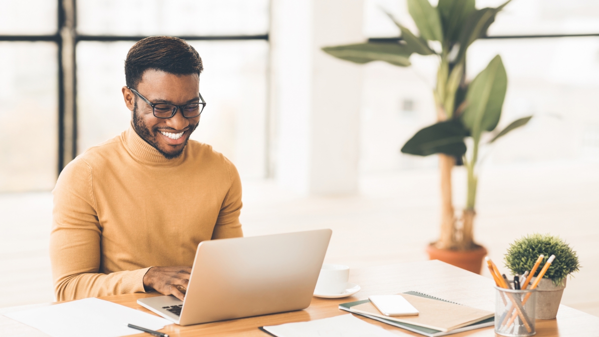 a happy black man smiling and working on laptop in home office.