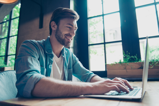 A happy, young, bearded man types on his laptop, representing using a best careers site