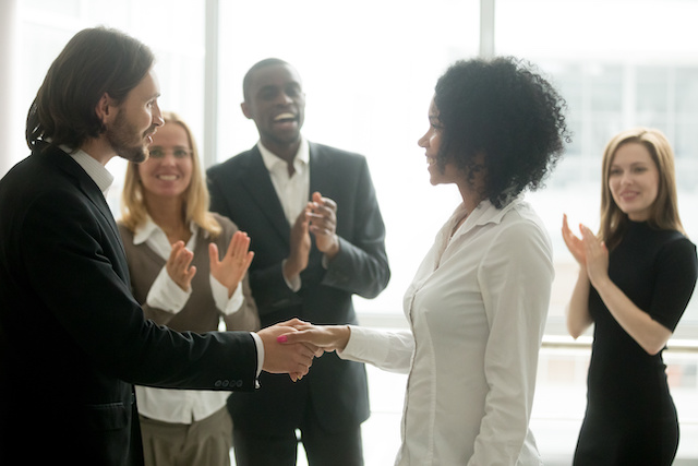 A male manager shakes the hand of his female employee in congratulations as three colleagues applaud her