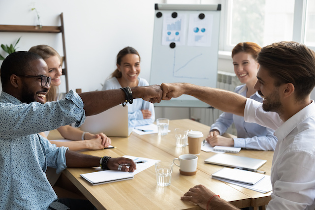 Two male colleagues fist bump across a meeting table as three female coworkers look on, smiling