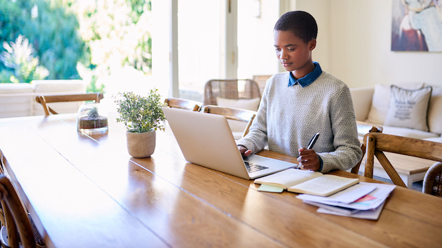 A young Black woman works on her laptop at her dining table at home