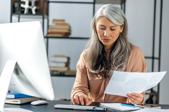 A gray-haired Asian HR director works through reports to determine the impacts of furloughs vs layoffs.