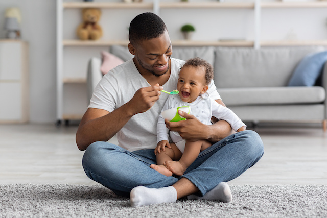 A Black father sits on a carpeted floor feeding his happy baby