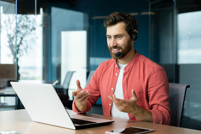 A bearded man in a red shirt has a video call on his laptop