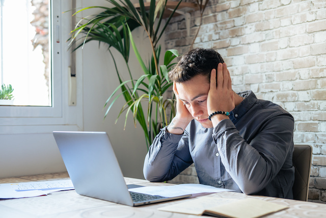 A male furloughed employee sits at his laptop with his head in his hands after learning he's been laid off.