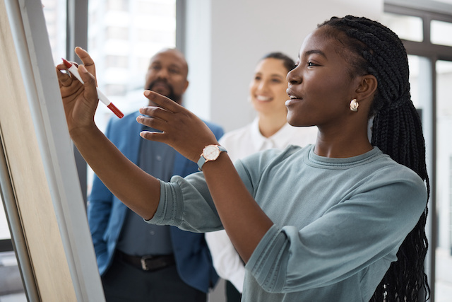 A young Black woman writes on a board during a learning session with two other colleagues