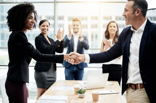 Executives from two companies shake hands among coworkers after finalizing a merger