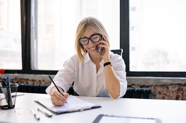 A blonde, female hiring manager wearing glasses speaks on the phone with a job candidate while taking notes.
