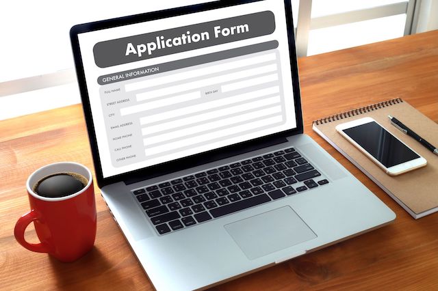 A laptop on a desk with a coffee mug, phone, pen and notebook displays a job application form