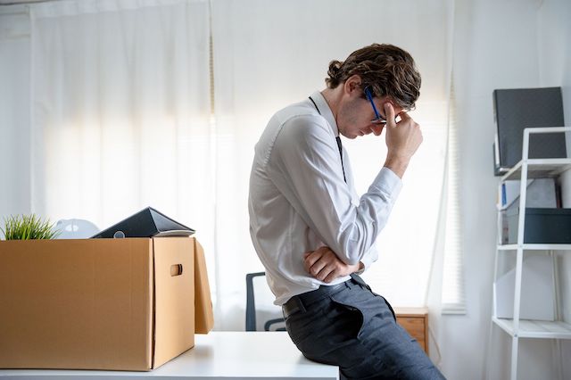 A terminated male employee leans against his desk, on which is a box of his belongings