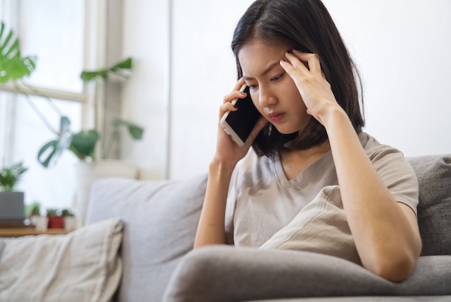 A distraught Asian woman sits on a couch talking on the phone