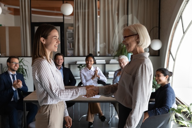 Established female leader congratulates young female employee in room full of colleagues