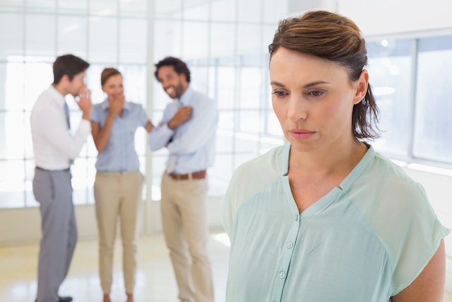 A woman looks unhappy with colleagues gossiping behind her back