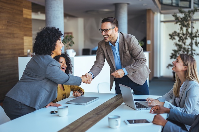 A male manager reaches across a table to shake the hand of a female client in a meeting with two other women