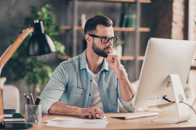 A bearded man participates in an online training from his office, watching intently