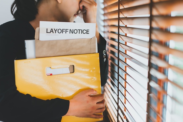 A female employee who has just been laid off stands near a window covered in blinds, while holding a box of her belongings