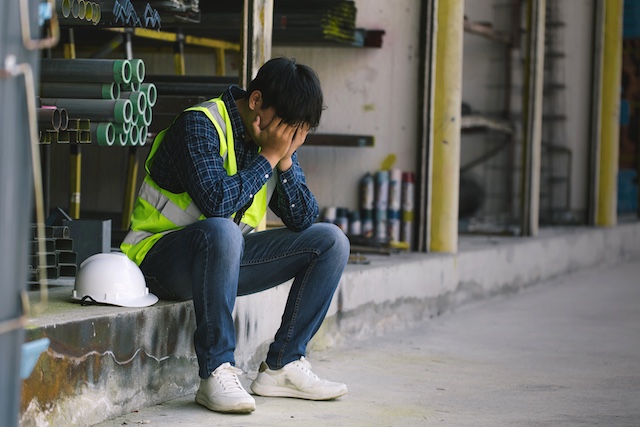 A male factory worker sits with his head in his hands, stressed about layoffs