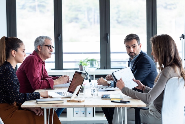 Four team members sit at a conference table for a meeting in a room with large windows