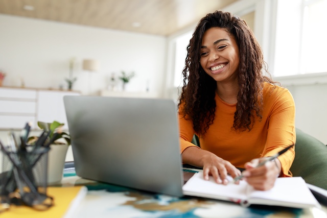 A young Black woman with long hair sits at a desk with her open laptop while watching a webinar