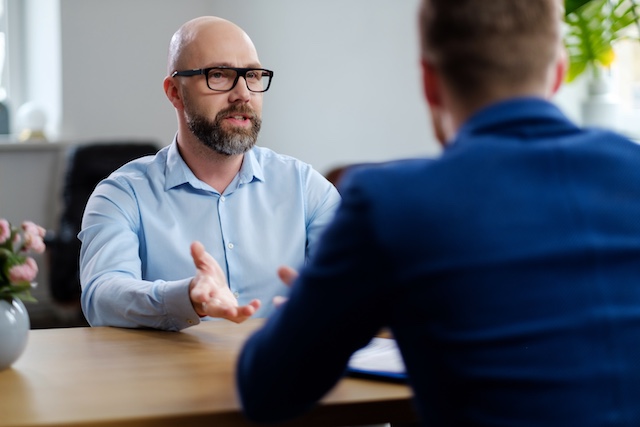 A middle-aged man with a beard and glasses is interviewed by a younger male manager