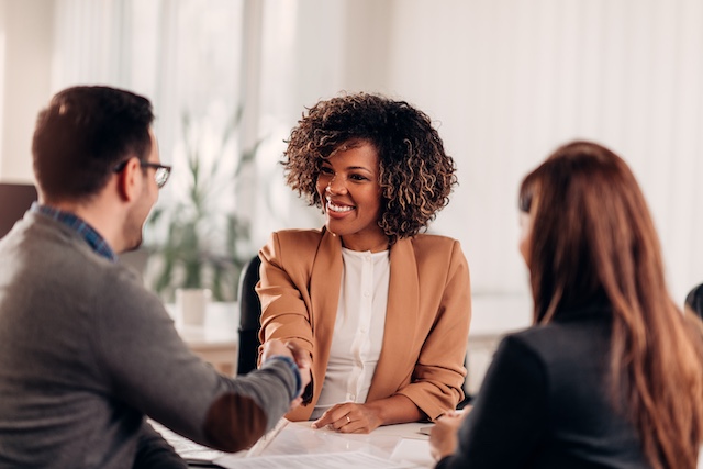A Black female executive shakes hands with a male executive who sits across from her with a woman after agreeing on executive compensation